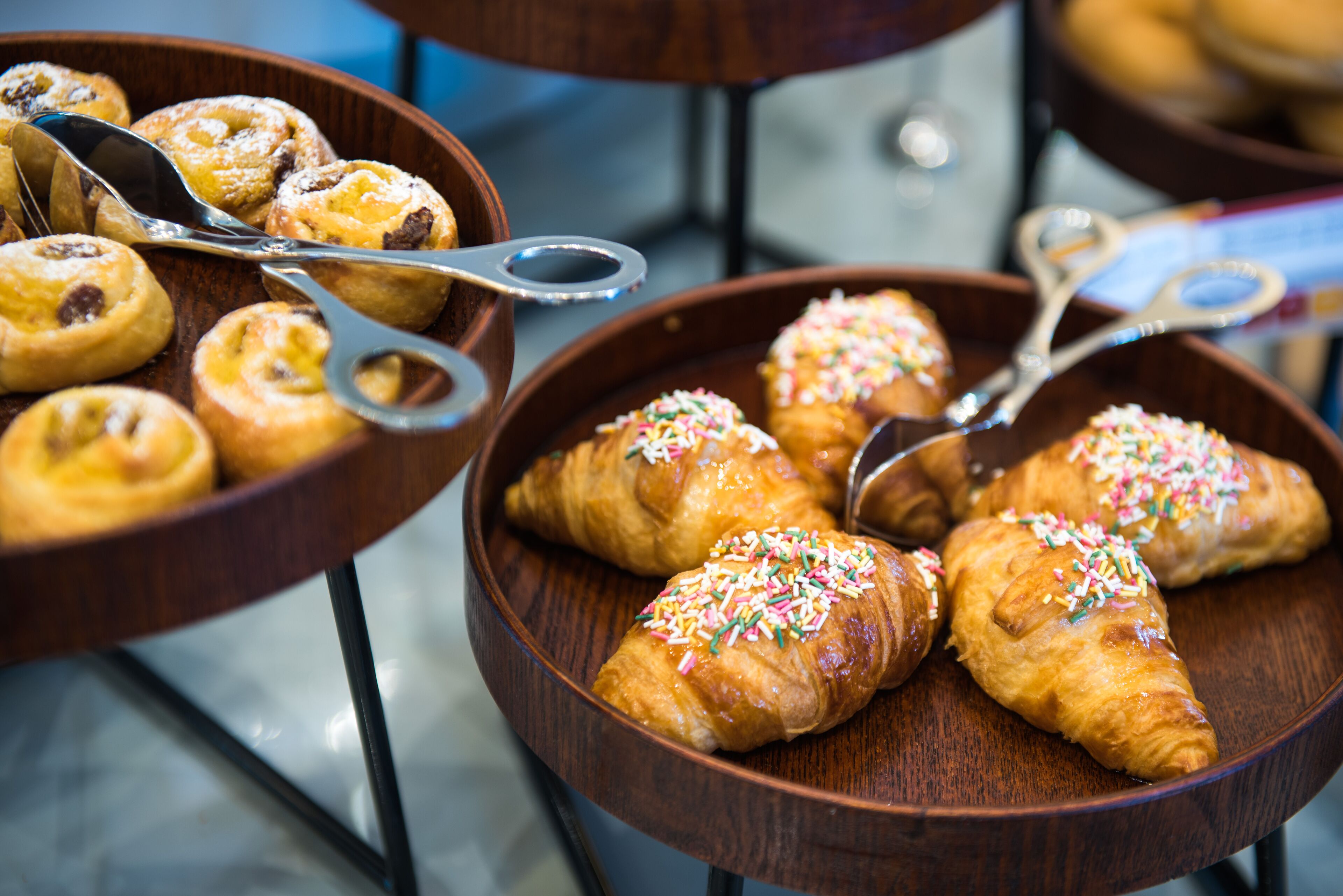 A variety of pastries in wooden bowls, including croissants and donuts, with serving utensils.