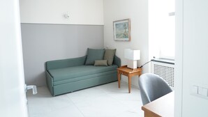 Een minimalistische woonkamer met een groene bank, een klein houten bijzettafeltje, een lamp en een ingelijst kunstwerk aan de muur.