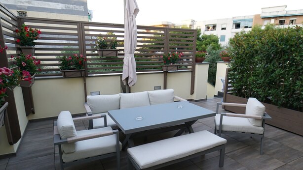 Een terras met een grijze tafel, witte fauteuils met kussens en een bankje. Op de achtergrond staan potplanten en een houten omheining.
