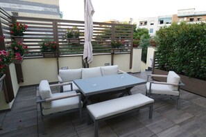 Une terrasse avec une table grise, des chaises blanches dotées de coussins et un banc. On aperçoit, à l’arrière-plan, des plantes en pot et une clôture en bois.