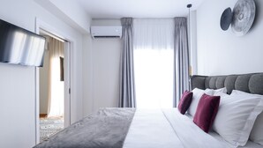 A modern hotel room with a large bed, grey and white bedding, a flat-screen TV mounted on the wall, and a ceiling fan. - Acropolis Grand Apartment (Athens)