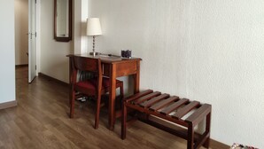 Una habitación con un escritorio de madera, una silla y un banco, un televisor montado en la pared y un espejo.