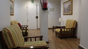 Un hall de hotel con sillones a rayas, un banco de madera, un extintor, una puerta con letrero de salida y un cuadro enmarcado en la pared.