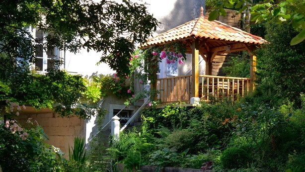 A wooden gazebo with a tiled roof surrounded by lush greenery and flowering plants.