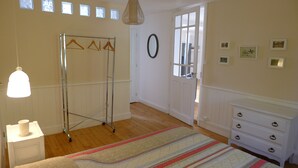 A room with a wooden floor, a bed with a striped blanket, a white dresser, a mirror, and a clothes drying rack.