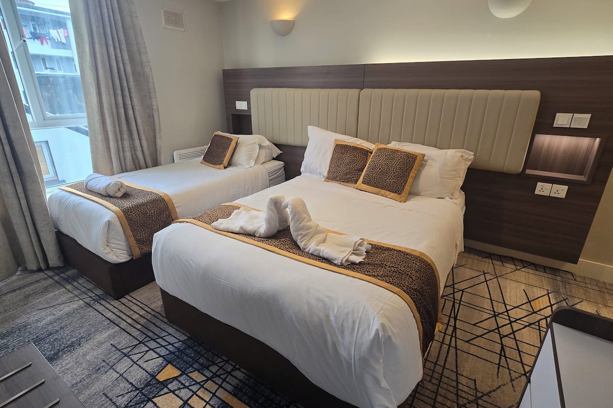 A hotel room with two beds, each with a headboard, white linens, and decorative pillows. There are wall-mounted light fixtures and a visible power outlet.