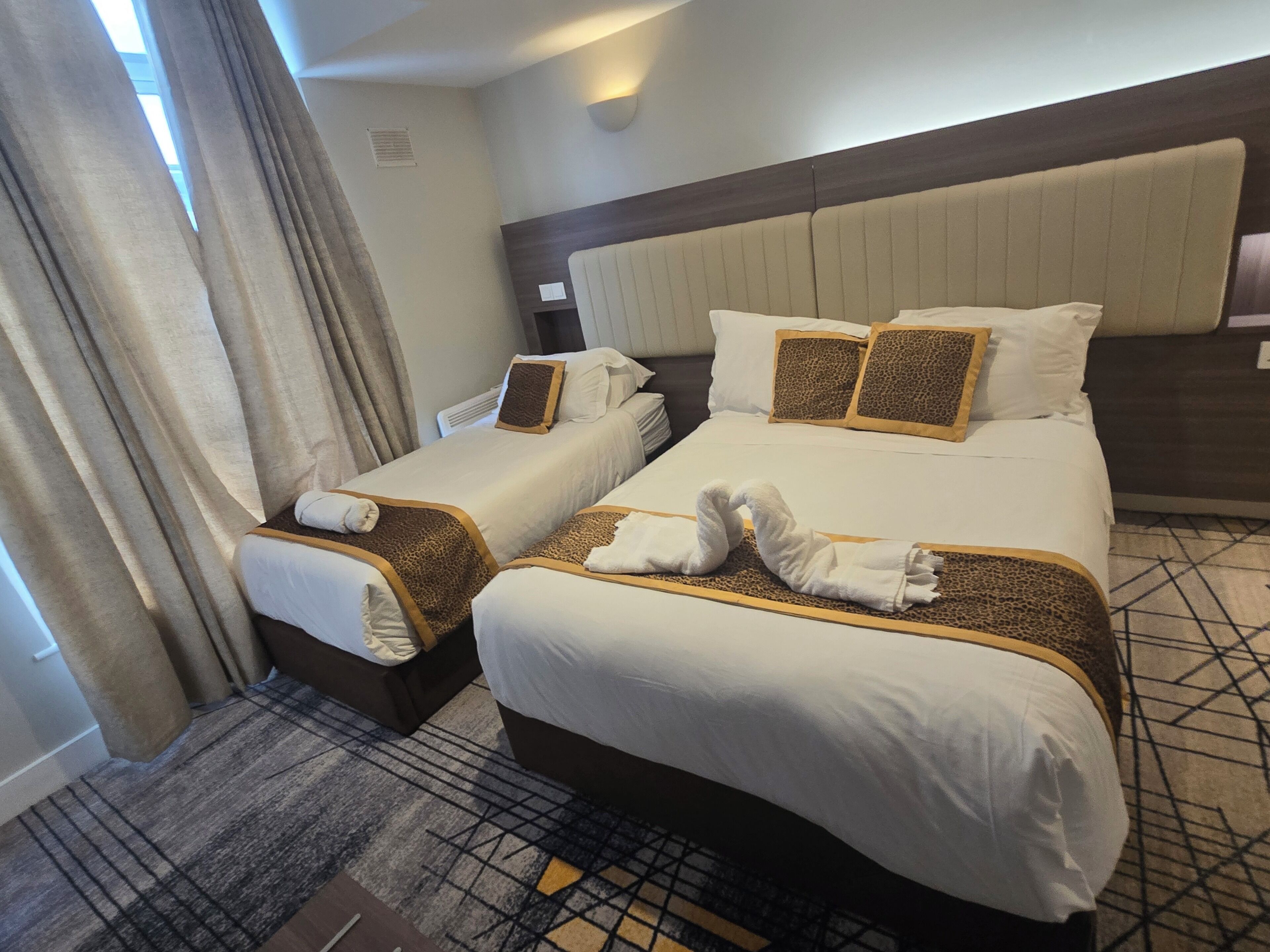 A hotel room with two beds, each with white linens and leopard print accents. There are towels arranged on the beds, and the room has a patterned carpet.