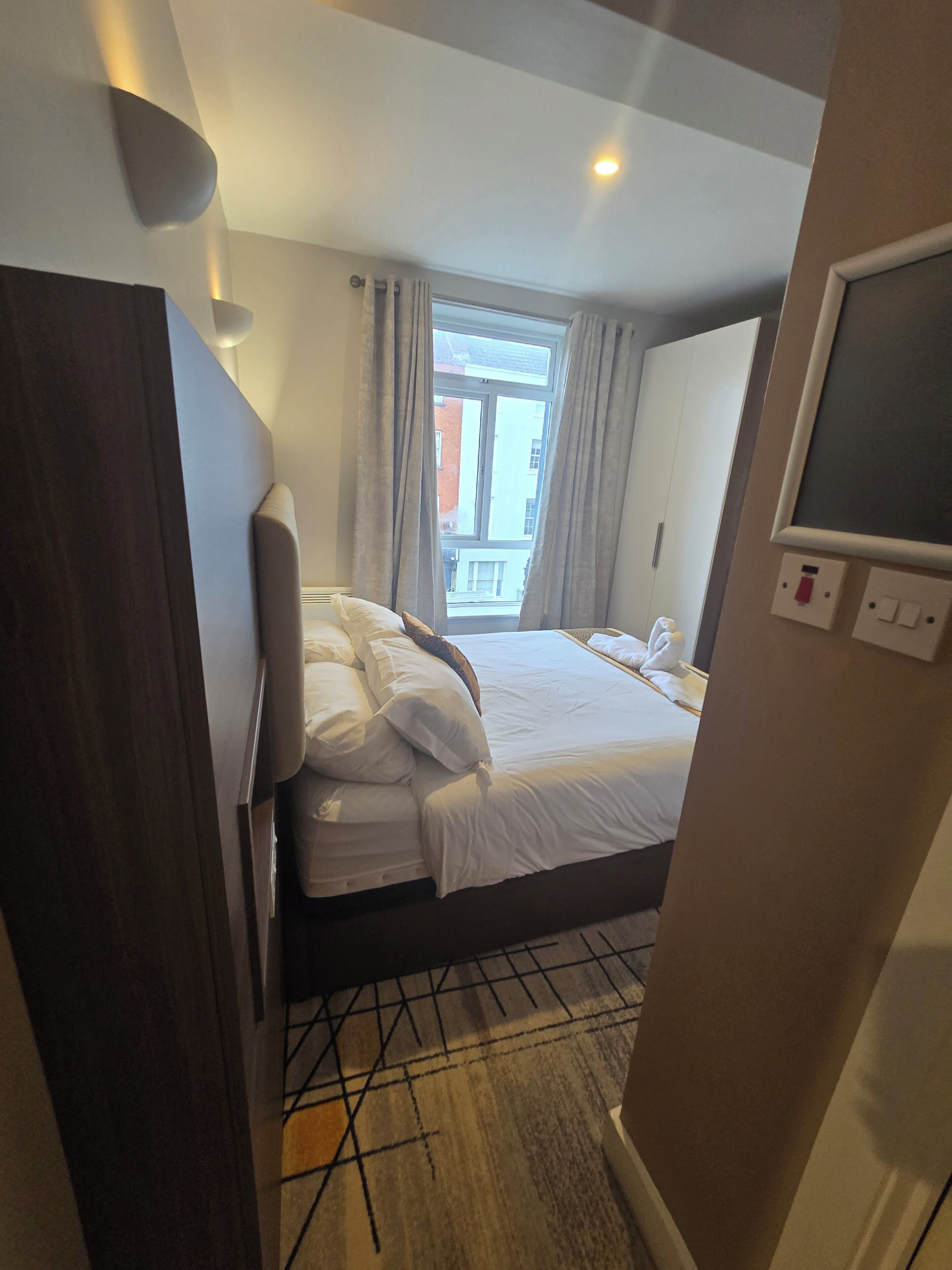 Ett hotellrum med en säng, en tv, ett fönster med gardiner och utsikt över byggnader utanför.