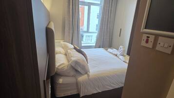 Ett hotellrum med en säng, en tv, ett fönster med gardiner och utsikt över byggnader utanför.