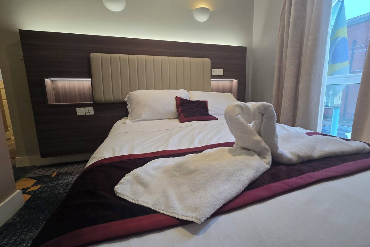 A hotel room with a bed, a headboard, a window with curtains, and a wall-mounted light fixture.