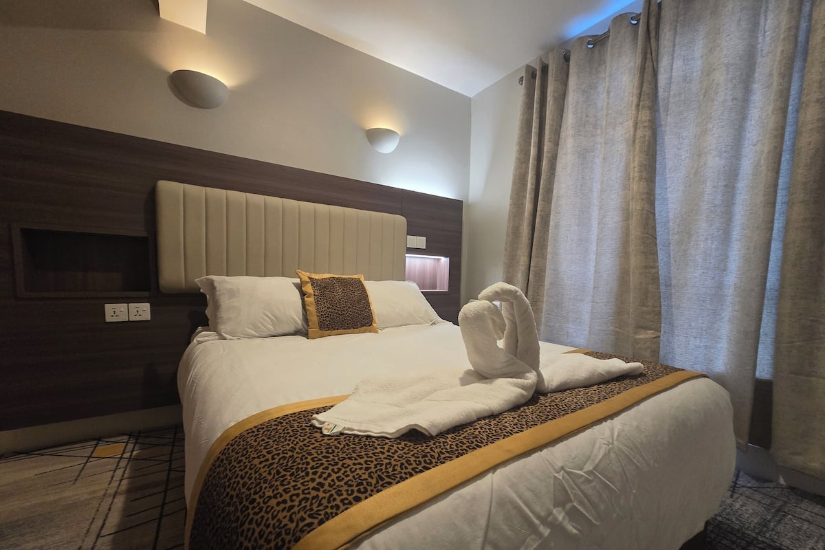 A hotel room with a large bed, a headboard, a wall-mounted light, and a window with curtains.