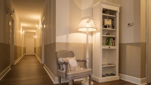 Un pasillo con suelo de madera, una silla con lámpara y una estantería.