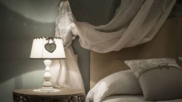 A bedroom with a bedside table, a lamp with a heart-shaped lampshade, a bed with white bedding, and a sheer white curtain.