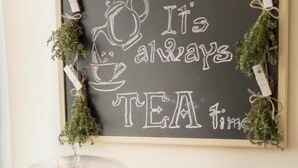 Una pizarra colgada en la pared con la frase 'It's always tea time' rodeada de follaje.
