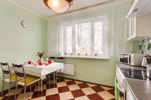 A dining area with a table set for two, a window with lace curtains, a microwave, and a stove. - Apartment Iris (Moscow)