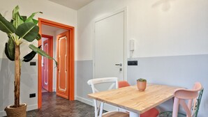 Huoneessa on puinen pöytä, valkoiset tuolit, ruukussa oleva kasvi ja ovi, jonka karmit ovat oranssit.