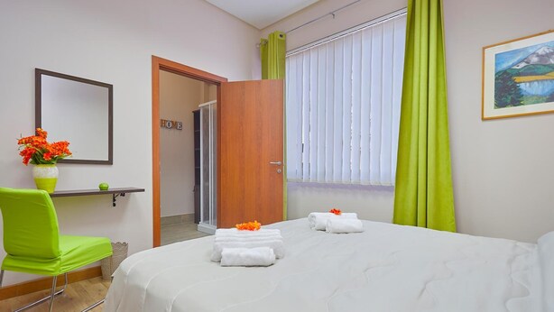 A hotel room with a bed, a green chair, a mirror, a painting, and a window with curtains. - Sturzo 44 (Palermo)