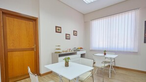 A room with a wooden door, white walls, a white table with chairs, a small shelf with decorative items, and a window with blinds. - Sturzo 44 (Palermo)