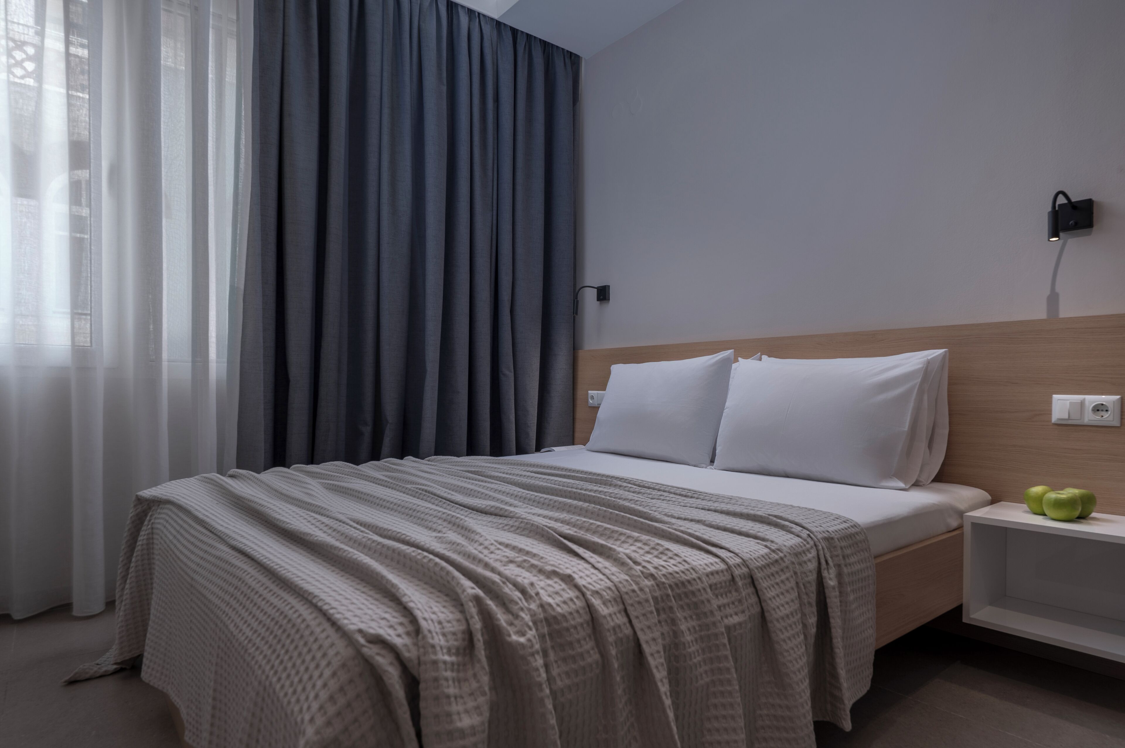 A hotel room with a bed, a nightstand, a wall socket, and a window with curtains.