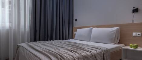 A hotel room with a bed, a nightstand, a wall socket, and a window with curtains.