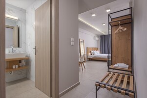 A modern hotel room with a bathroom, a bed, a wooden bench, and a hanging clothes rack.