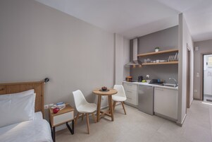 A compact hotel room with a bed, a small dining table with chairs, a kitchenette with a sink and cabinets, and a window.