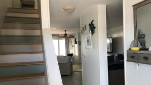 Una escalera conduce a una sala de estar con ventilador de techo, una cocina con refrigerador y una mesa de comedor.