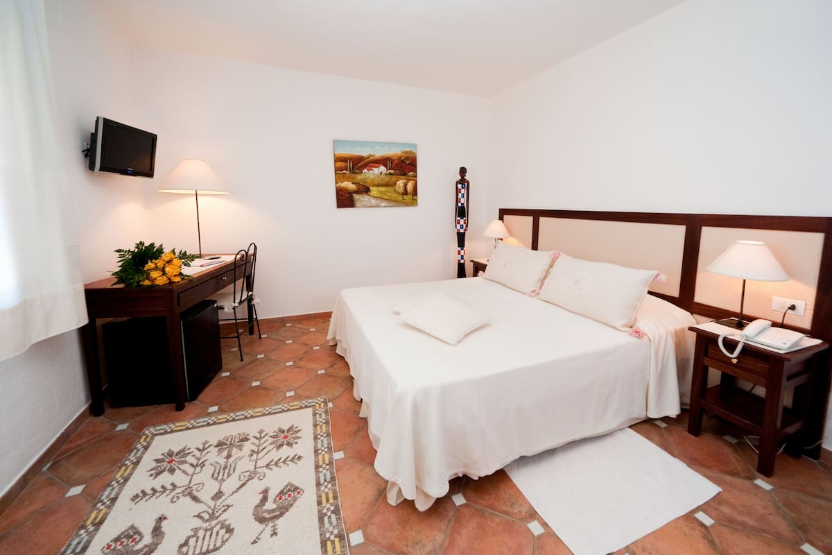 A hotel room with a bed, bedside tables, a desk with a lamp, a television, and a painting on the wall.