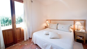 A bedroom with a large bed, two bedside tables with lamps, a wooden door, and a view of greenery outside. - S'Arenada Hotel (Villasimius)