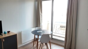 Una habitació petita i lluminosa amb una taula rodona i dues cadires, un armari i una finestra gran amb cortines.