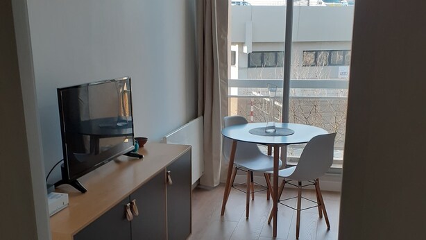 Habitació moderna amb televisió a un moble, taula petita amb cadires i finestra gran que mostra edificis.