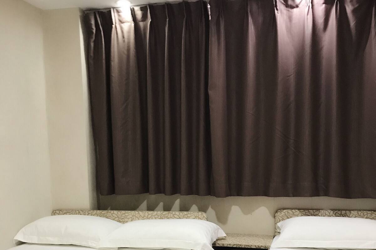 A hotel room with two single beds, a nightstand, and a large window with dark curtains.
