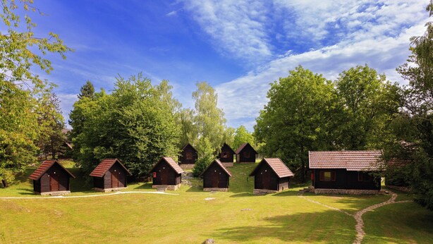 A row of wooden cabins with stone foundations, surrounded by lush greenery and trees, under a clear blue sky. - Kamp Korana (Rakovica)