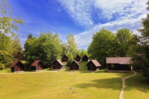A row of wooden cabins with stone foundations, surrounded by lush greenery and trees, under a clear blue sky. - Kamp Korana (Rakovica)