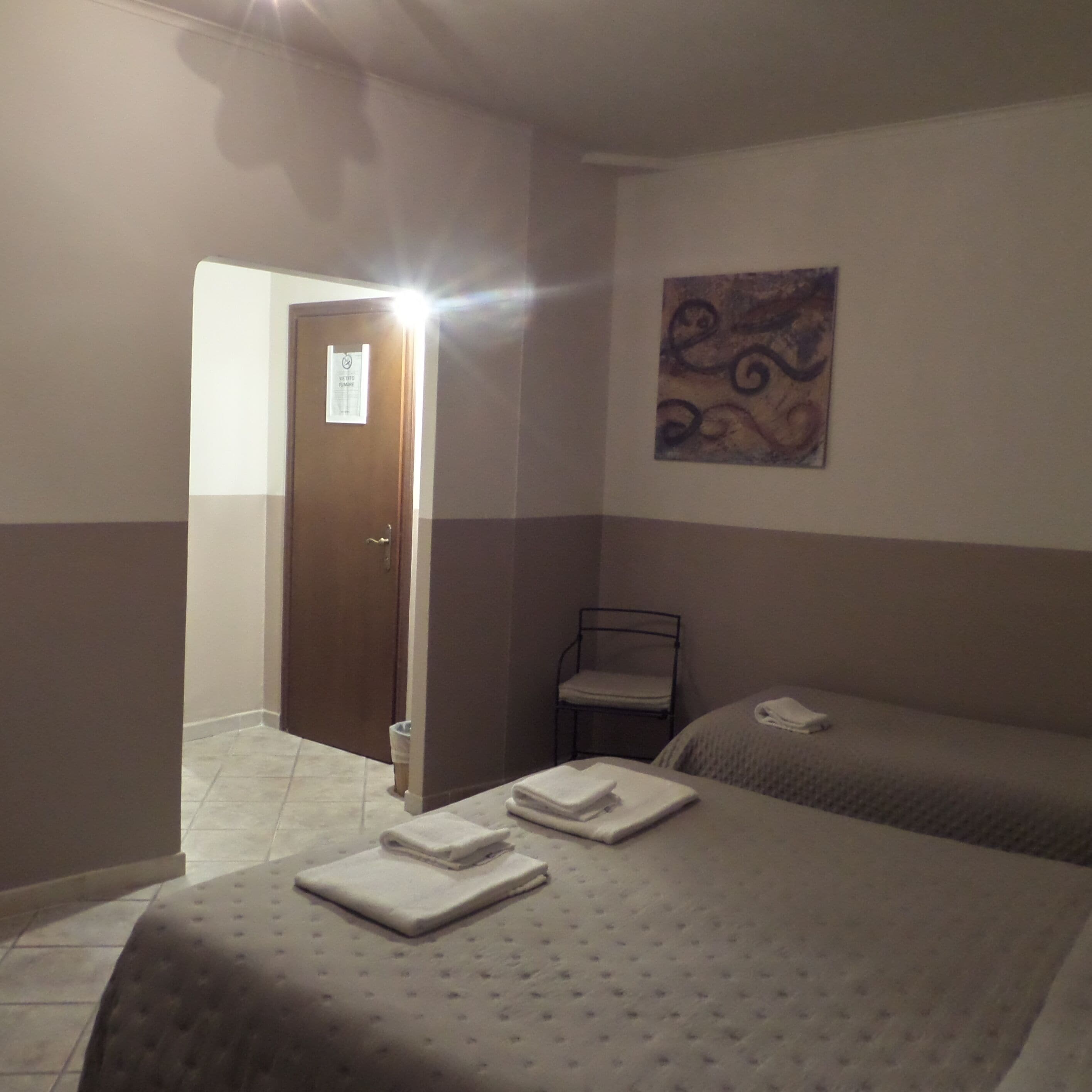 Una camera d'albergo con due letti, una sedia, un quadro appeso al muro e una porta con un cartello.
