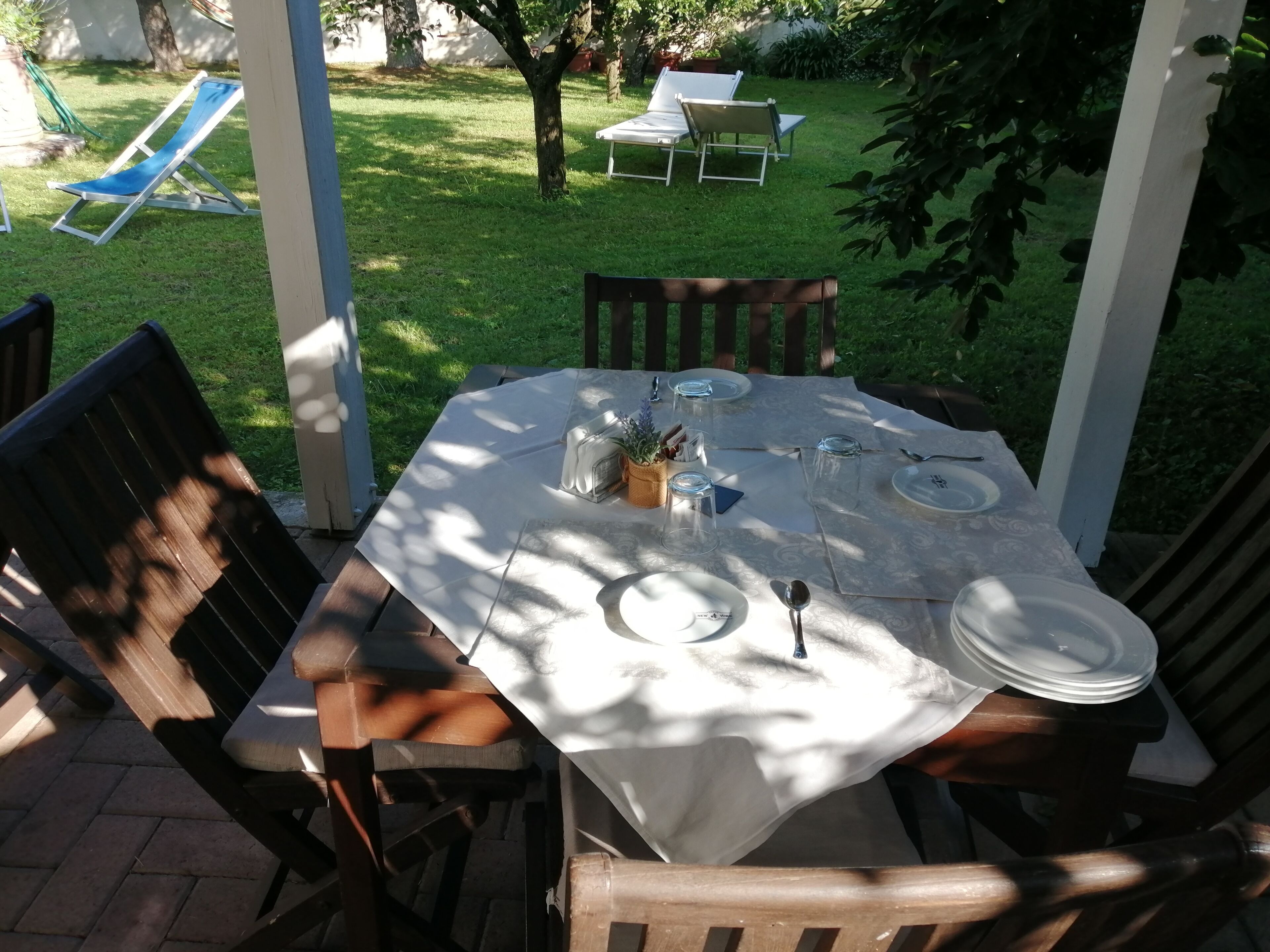 Un patio al aire libre con una mesa lista para cuatro personas, rodeada de sillas de madera, en un jardín soleado.