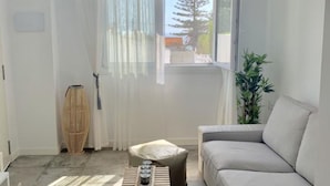 En moderne stue med en grå sofa, et lille sofabord, en fletstol og et stort vindue med lette gardiner.