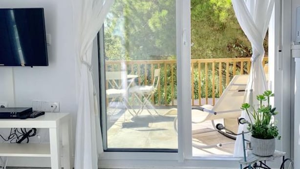Et værelse med et fjernsyn på væggen, et skrivebord med en computer, og et stort vindue med udsigt til en terrasse med udendørs møbler.