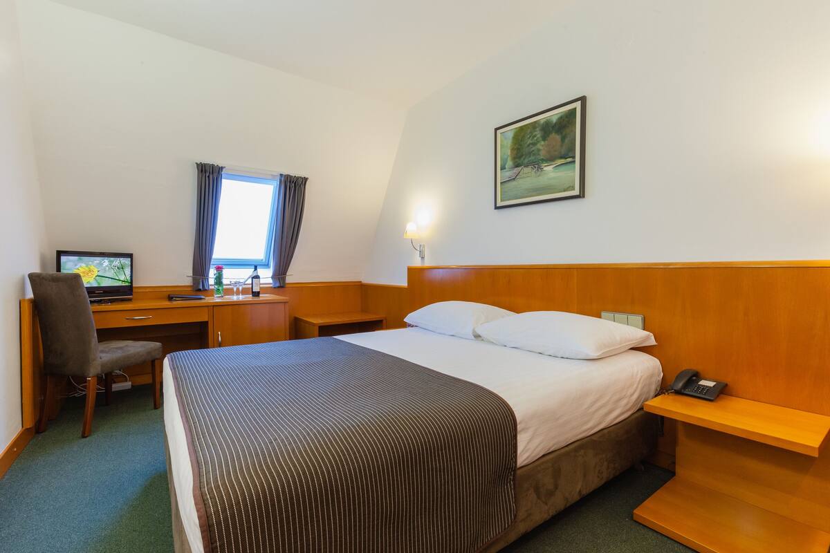 A hotel room with a bed, desk, chair, and a painting on the wall.