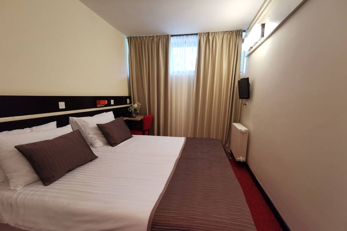 A hotel room with a large bed, a desk, a chair, a radiator, and a TV on the wall.