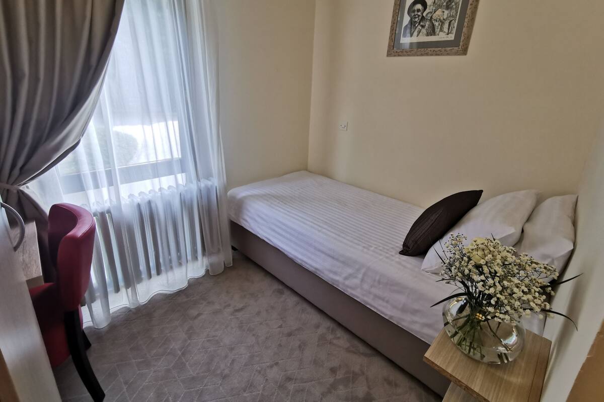 A small, single-bed room with a window, a chair, a bedside table with a vase of flowers, and a framed picture on the wall.