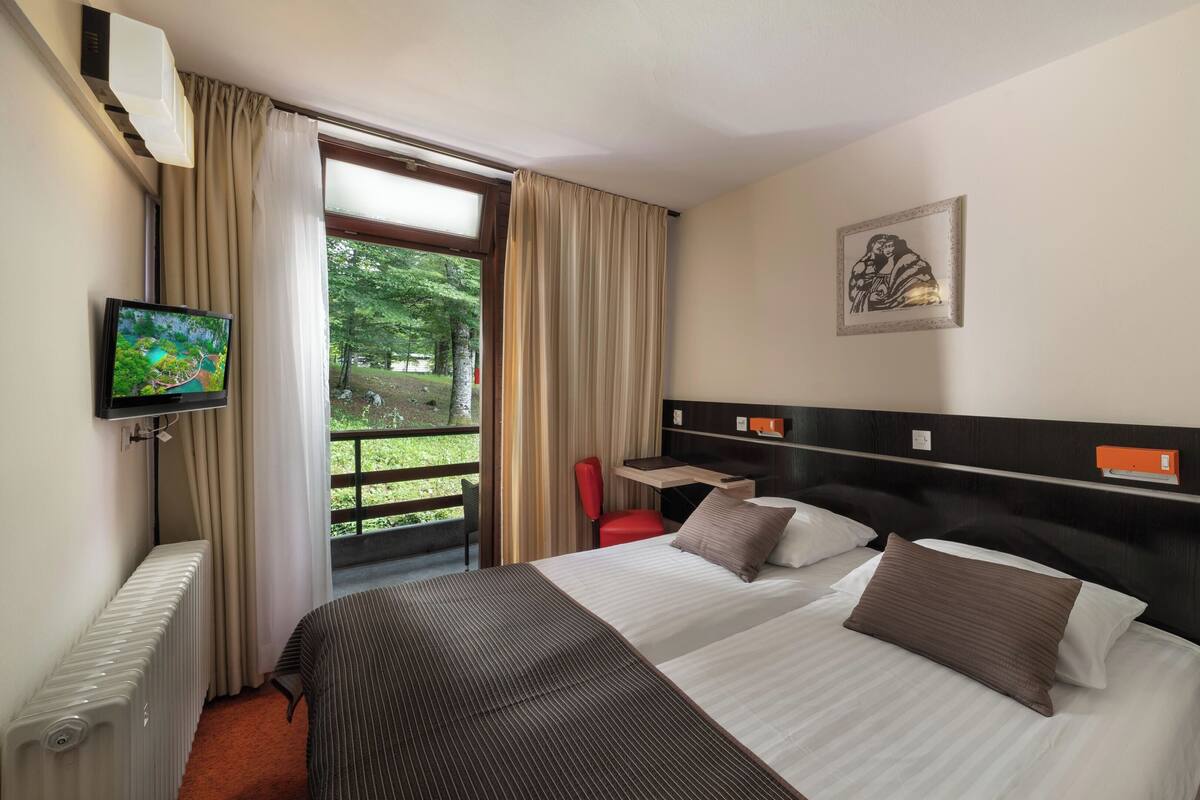 A hotel room with a bed, a television, a window with curtains, a radiator, and a painting on the wall.