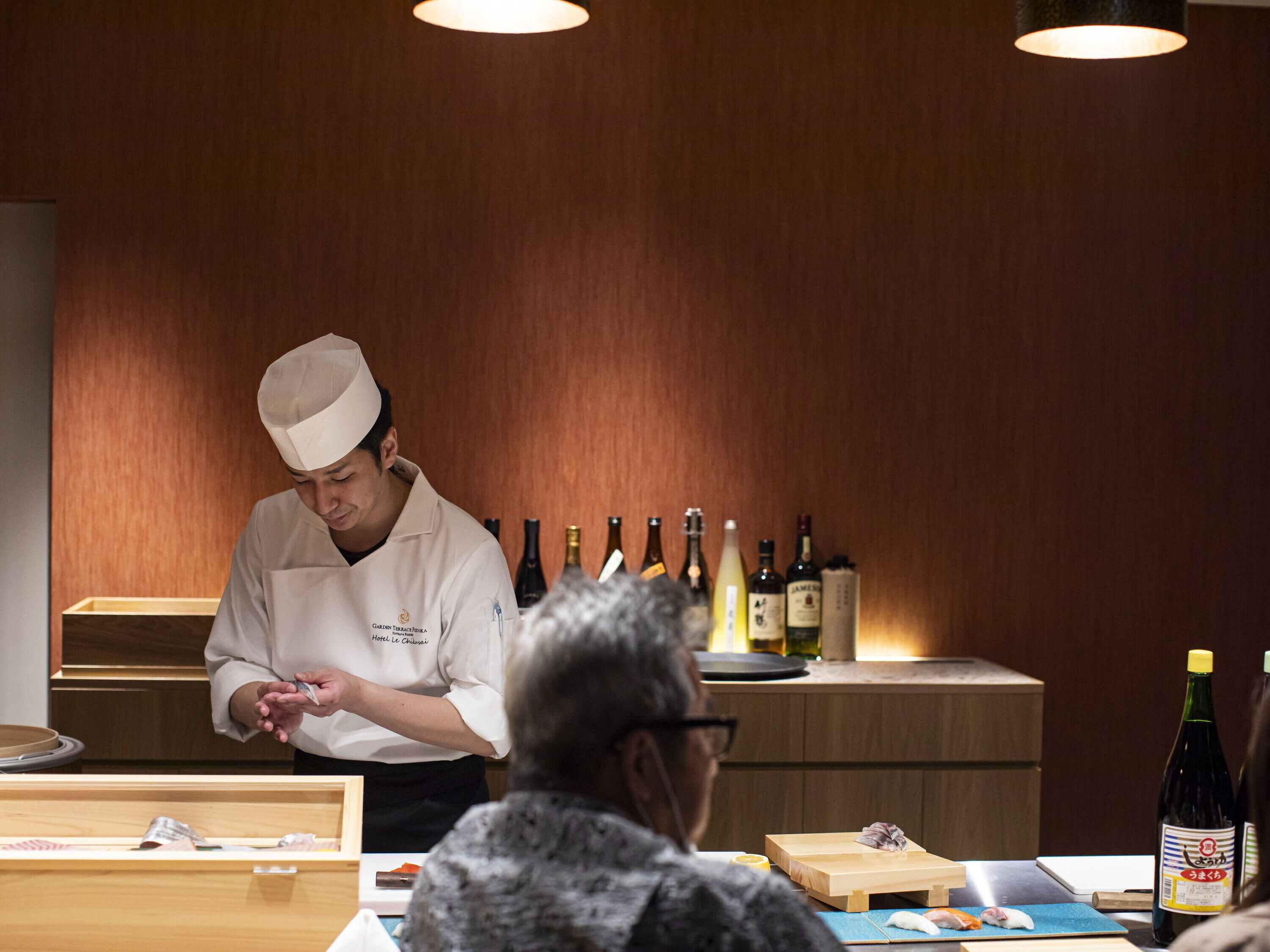 Un chef preparando comida en un restaurante con paredes de madera y una barra que exhibe varias botellas.