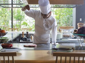 A chef in a white uniform preparing food on a cutting board in a well-lit kitchen with a view of greenery outside. - Garden Terrace Fukuoka Hotel & Resort (Fukuoka)