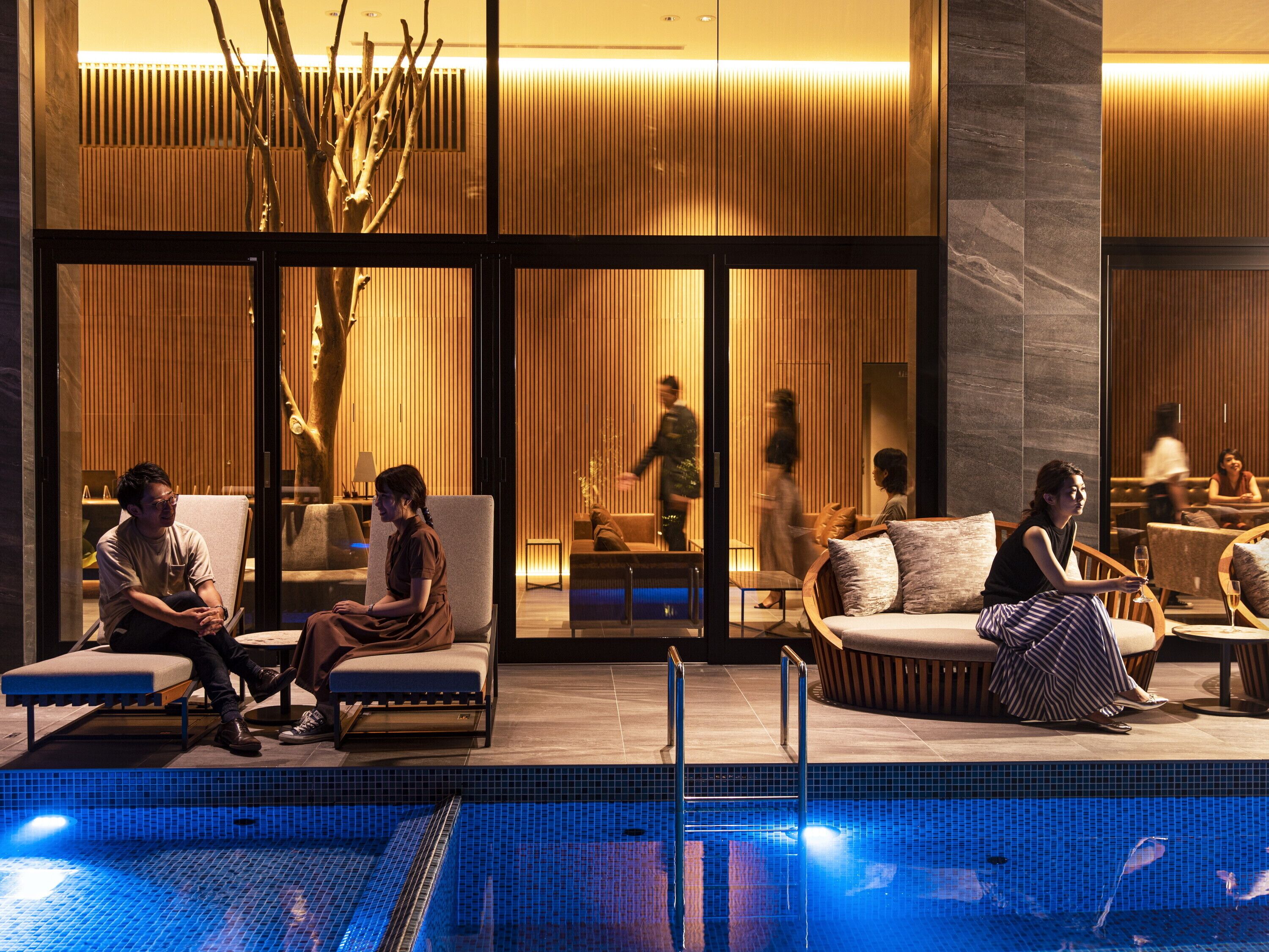 A modern hotel lobby with a pool area, seating arrangements, and a view of the interior through large glass doors.