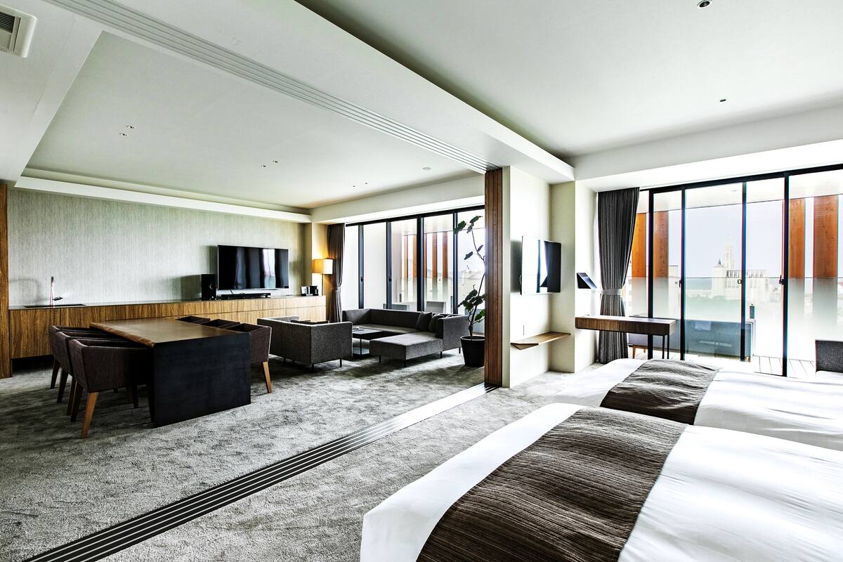 A modern hotel room with a large bed, a sofa, a dining table, and a TV. The room has large windows with a view of a building.