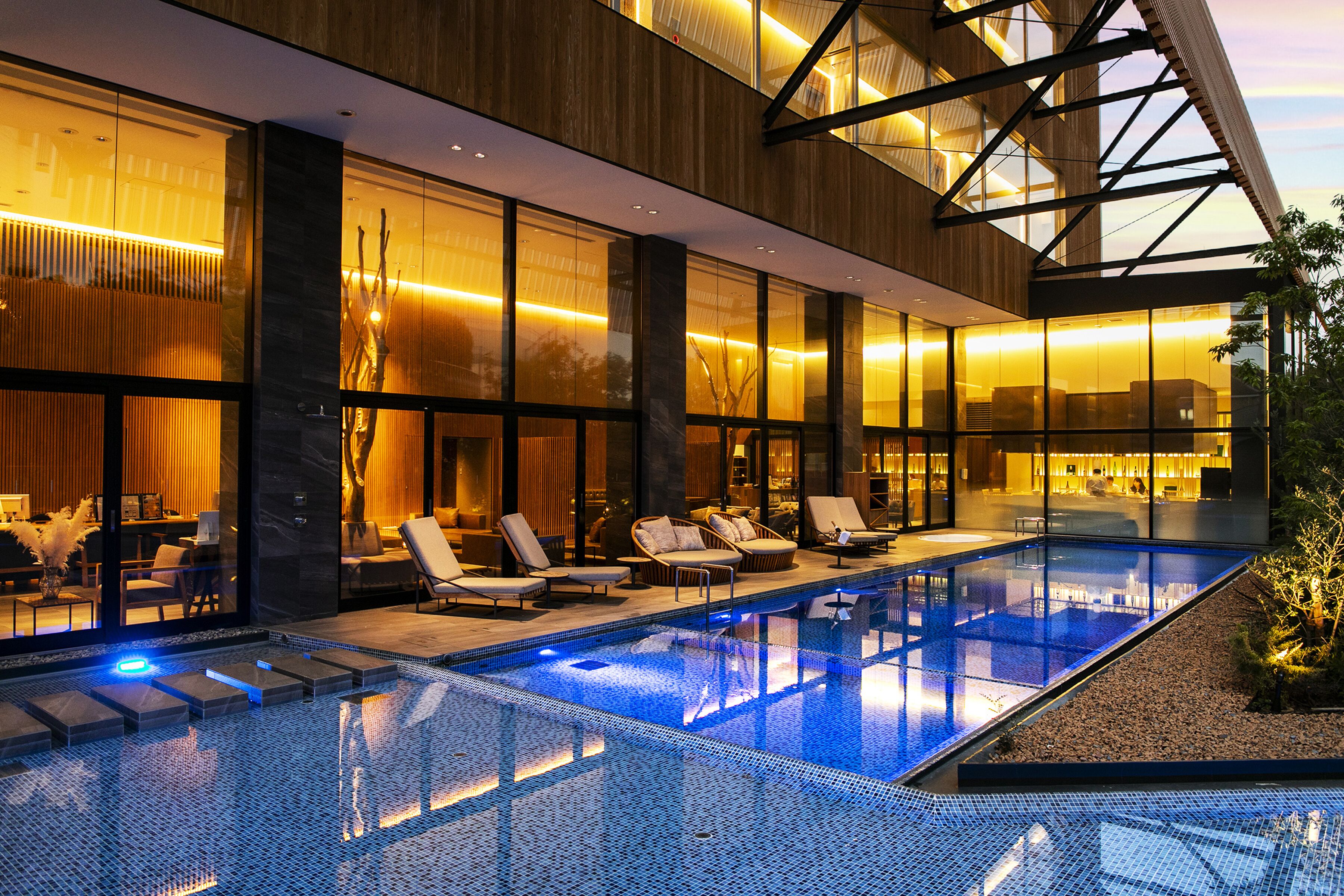 A modern hotel with a glass-enclosed infinity pool, lounge chairs, and a well-lit interior.