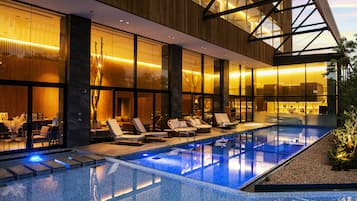 A modern hotel with a glass-enclosed infinity pool, lounge chairs, and a well-lit interior.