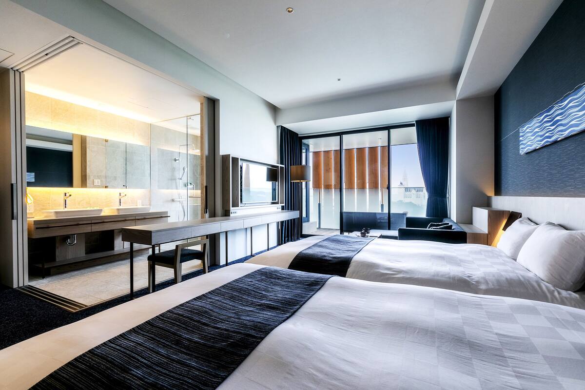 A modern hotel room with two beds, a large mirror, a desk, and a bathroom with a vanity.