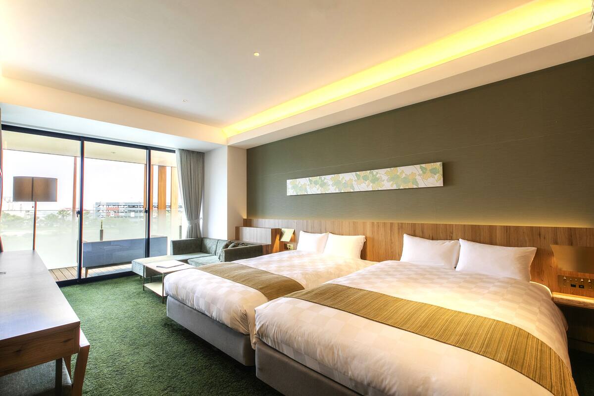 A modern hotel room with two beds, a dining area, and a large window offering a view.